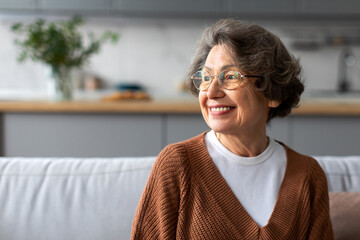 Portrait of happy senior woman relaxing on comfortable couch at home in living room and looking away at free space, enjoying domestic pastime