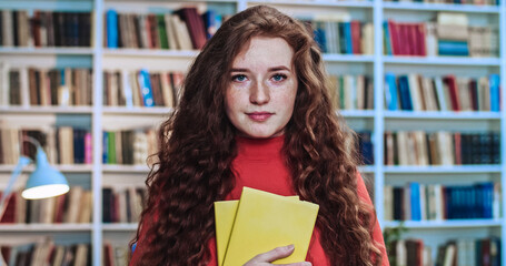 Portrait of cute red head sweet girl with long curly natural hair and yellow books studying in...