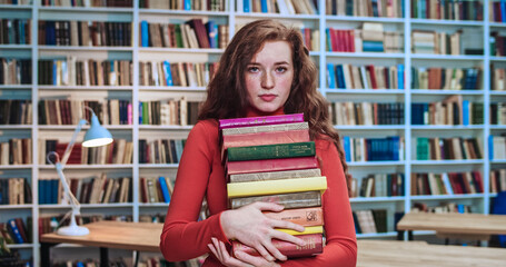 Close-up portrait of serious red head student with long curly natural hair and freckles looking into camera while keeping a lot of books in library. Bookcase in background.