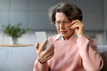 Confused senior woman in eyeglasses squinting eyes reading message on cellphone, having ophtalmic issue problems with vision sitting on couch - obrazy, fototapety, plakaty