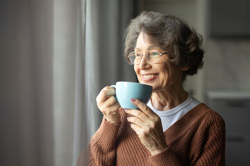 Happy senior woman drinking coffee looking out window at home, enjoying domestic rest, standing with cup in hands, looking aside and smiling