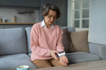 Caucasian senior woman measuring blood pressure, sitting on couch at home in living room