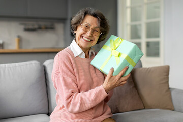 Joyful senior woman holding wrapped gift box near face and smiling, sitting on sofa at home,...