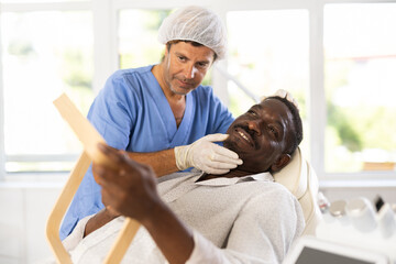 Experienced male cosmetologist examines facial skin of African-American man patient and advises on...