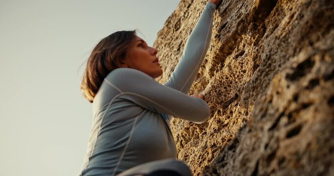 Shot close up, shot from below, a blonde girl in a blue jacket climbs a steep rock on which the rays of the sun fall. blonde girl climber climbs up a steep rocky rock and looks for where to put her