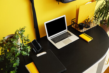 Modern laptop, mobile phone and stationery on dark table near color wall