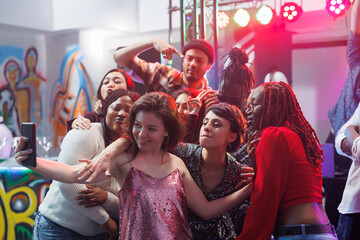 Group of friends posing for selfie on crowded nightclub dancefloor while clubbing together. Young carefree people taking photo on smartphone while enjoying discotheque event