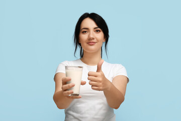 Pretty young woman with glass of milk showing thumb-up on blue background