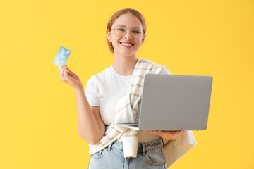Young woman with credit card and laptop on yellow background - obrazy, fototapety, plakaty