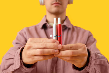 Young man with electronic cigarettes on yellow background, closeup