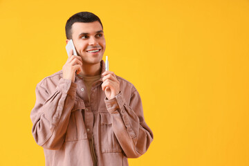 Young man with electronic cigarette talking by phone on yellow background