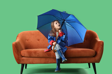 Shocked little girl with umbrella sitting on sofa against green background