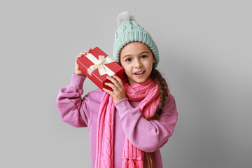 Cute little girl in warm hat and scarf with Christmas gift on grey background