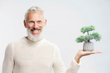 Smiling gray haired bearded man, gardener  holding bonsai plant looking at camera isolated on white background 