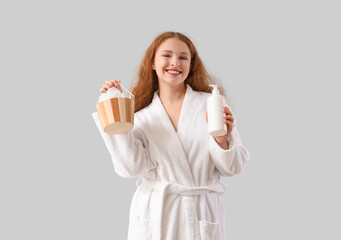 Young woman in bathrobe with shower supplies on light background
