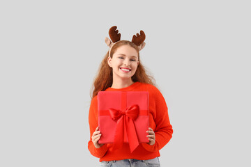 Young woman in reindeer horns with Christmas gift box on light background