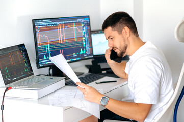 Busy caucasian male crypto trader sitting at home at his work desk in front of monitors with graphs, talking on the smart phone with colleague, discussing the prospects of cryptocurrency, profit