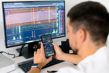 Crypto trader sits at his workplace in front of a monitor with charts holds smartphone in hands with trading data index chart graph on the screen, mobile phone app analytics for cryptocurrency market