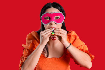 Happy young woman with carnival mask blowing in whistle on red background