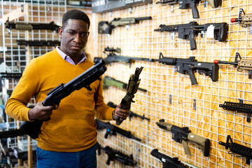 African-american man choosing assault rifle and shotgun in airsoft shop.