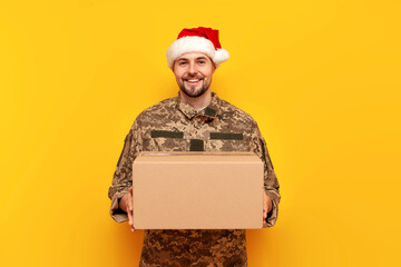 Ukrainian soldier in pixelated uniform and santa claus hat holding a box on a yellow isolated...