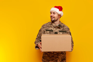 Ukrainian soldier in a pixelated uniform and santa claus hat holds a box on a yellow isolated background