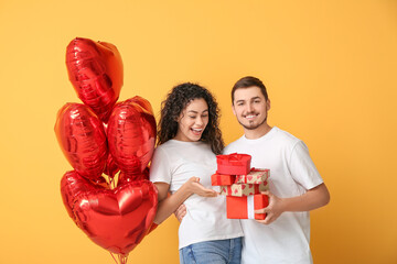 Young couple with gifts and air balloons in shape of heart for Valentine's day on yellow background