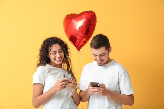 Young couple with mobile phones and air balloon in shape of heart for Valentine's day on yellow background