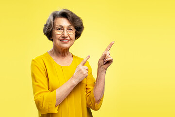 Beautiful smiling senior woman pointing fingers on copy space looking at camera isolated on yellow...