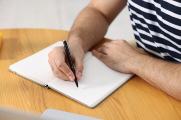 Young man writing in notebook at wooden table, closeup