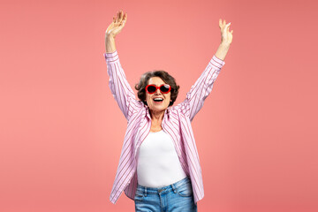 Overjoyed attractive senior woman, funny grandmother wearing red eyeglasses, stylish stripped shirt dancing, celebration success isolated on pink background. Emotions, positive lifestyle concept