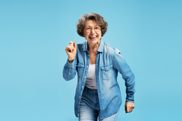 Smiling confident senior woman, happy active grandmother wearing stylish denim jacket, eyeglasses dancing isolated on blue background. Positive lifestyle concept