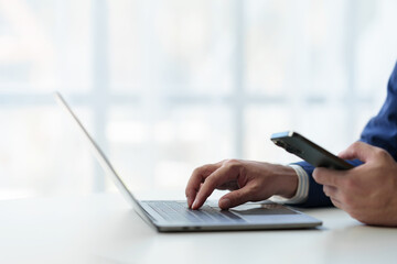 Businessman holds smartphone, checks email, checks job details on Laptop computer on white desk to...