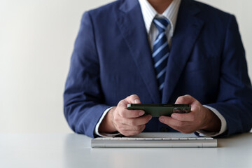 Young Asian businessman holds cell phone, points at smartphone, checks email, plays online shopping game. Typing work on a pc computer keyboard on a white desk in a corporate office.