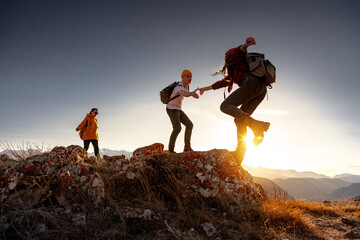 Group of young hikers with backpacks walks with backpacks and helps each other in climbing in sunset mountains - obrazy, fototapety, plakaty