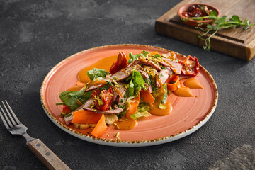 A beautifully plated duck salad with vibrant carrot puree, served on a rustic peach-colored plate against a black textured backdrop