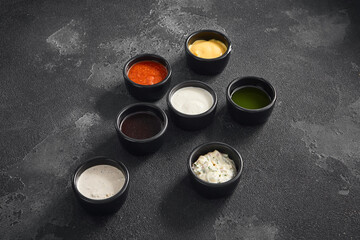A variety of dipping sauces presented in black ramekins against a textured dark backdrop, perfect...
