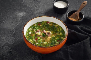 Traditional okroshka with beef served in an orange bowl on a dark slate background with sour cream and spices nearby
