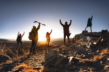 Big group of young hikers with backpacks are standing with open arms in winner poses at sunset...
