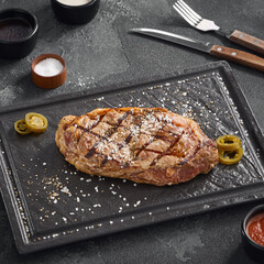 A juicy ribeye steak with grill marks, seasoned with sea salt and cracked pepper, served on a black stone plate with a side of jalapenos