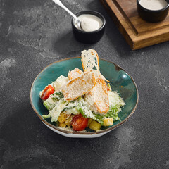 Classic Caesar salad with grilled chicken, croutons, parmesan, and tomatoes, served in a teal bowl on a dark textured surface