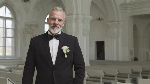Medium portrait of handsome Caucasian grey haired bearded groom in elegant suit and buttonhole posing for camera in church before wedding ceremony