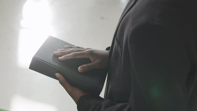 Cropped shot of unrecognizable Black male priest holding Bible while standing in sunlit church