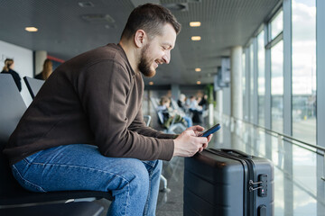 portrait of cheerful man relaxing at airport with bag and mobile phone, guy waiting for flight using mobile phone communicating in social media, searching information online. 