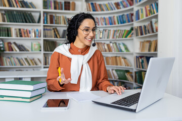 Smiling Indian businesswoman in headset sits at laptop in room, communicates with clients online, works in call center, looks at screen.