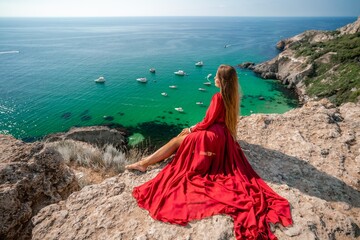 Woman red dress sea. Happy woman in a red dress and white bikini sitting on a rocky outcrop, gazing...