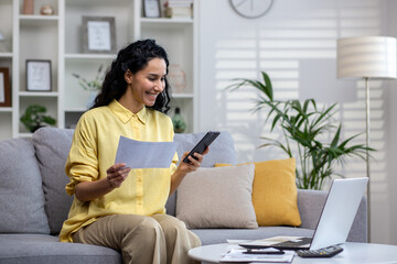 Happy hispanic woman working in home office sitting on sofa in front of laptop, holding phone and...