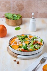 Green salad with persimmon and boiled chicken in a white plate on a light concrete background. Persimmon recipes.
