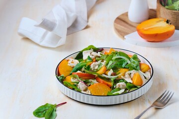 Green salad with persimmon and boiled chicken in a white plate on a light concrete background. Persimmon recipes.