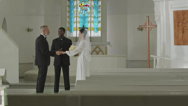 Wide full length slowmo shot of young African American male officiant declaring mature Caucasian man and woman husband and wife during private church wedding ceremony